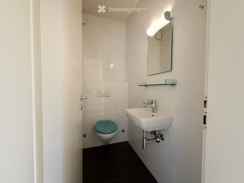 Modern bathroom with white tiles, black floor, white sink with chrome faucet, toilet with turquoise seat, mirror with built-in light, and a glass shelf above the sink.
