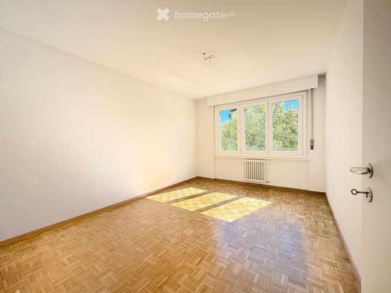 Empty room with wooden herringbone flooring, white walls, and large windows showing greenery outside. No furniture or equipment present.