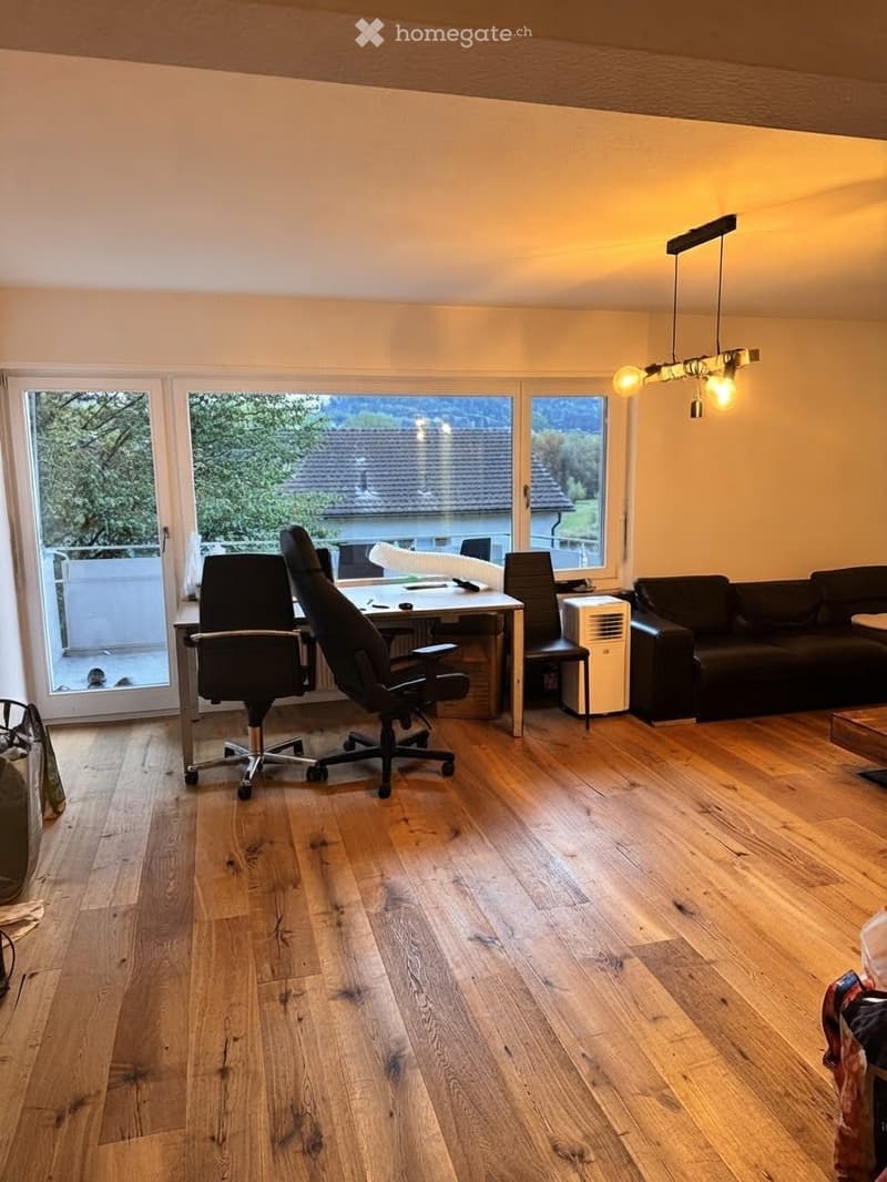 Bright living room with wooden flooring, large windows, a sofa, office chair, desk with laptop, and an air conditioner unit.