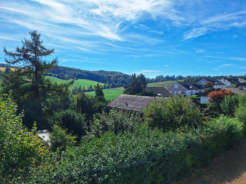 A peaceful residential area with houses surrounded by lush greenery and trees, set against a backdrop of rolling hills and a clear blue sky.