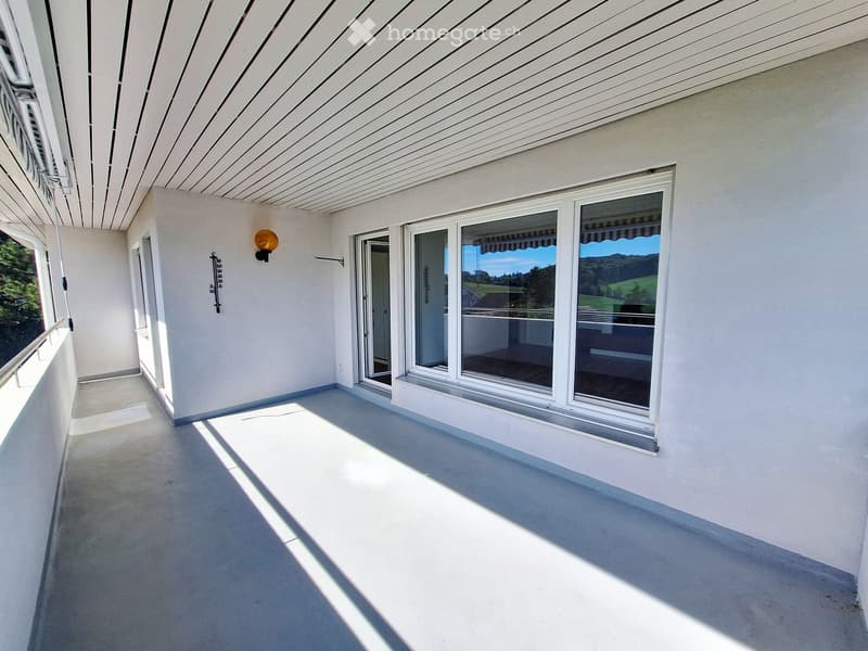 Spacious balcony with white walls, large windows, and a view of the landscape. Features a modern ceiling design and a weather station mounted on the wall.