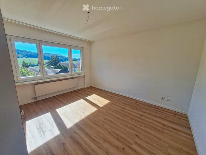 Empty room with wooden flooring, large windows offering a scenic view, and a radiator below the windows.