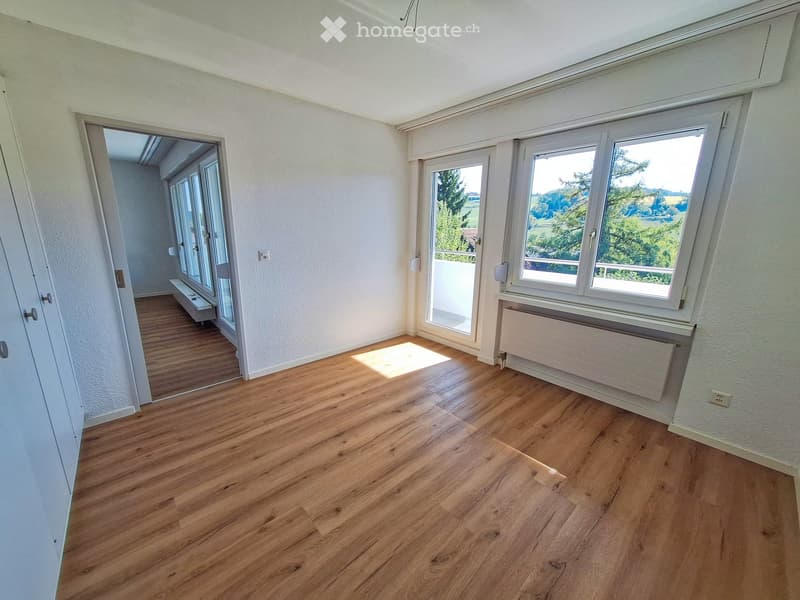 Empty room with wooden flooring, white walls, large windows, and a sliding glass door leading to a balcony.