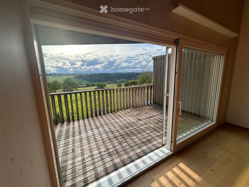 Open sliding glass door leads to a wooden balcony with a railing, overlooking a green field and distant landscape.