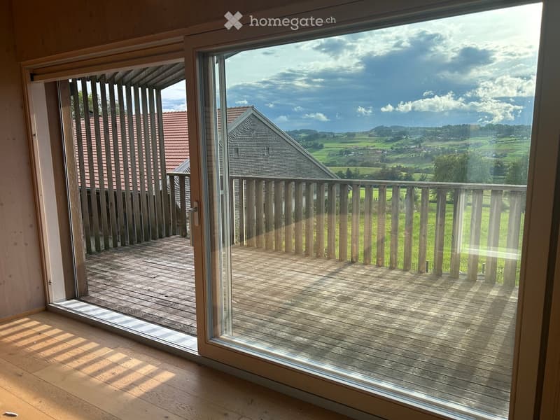 Open sliding glass doors lead to a wooden deck with a railing, overlooking a scenic countryside with fields and a distant house.
