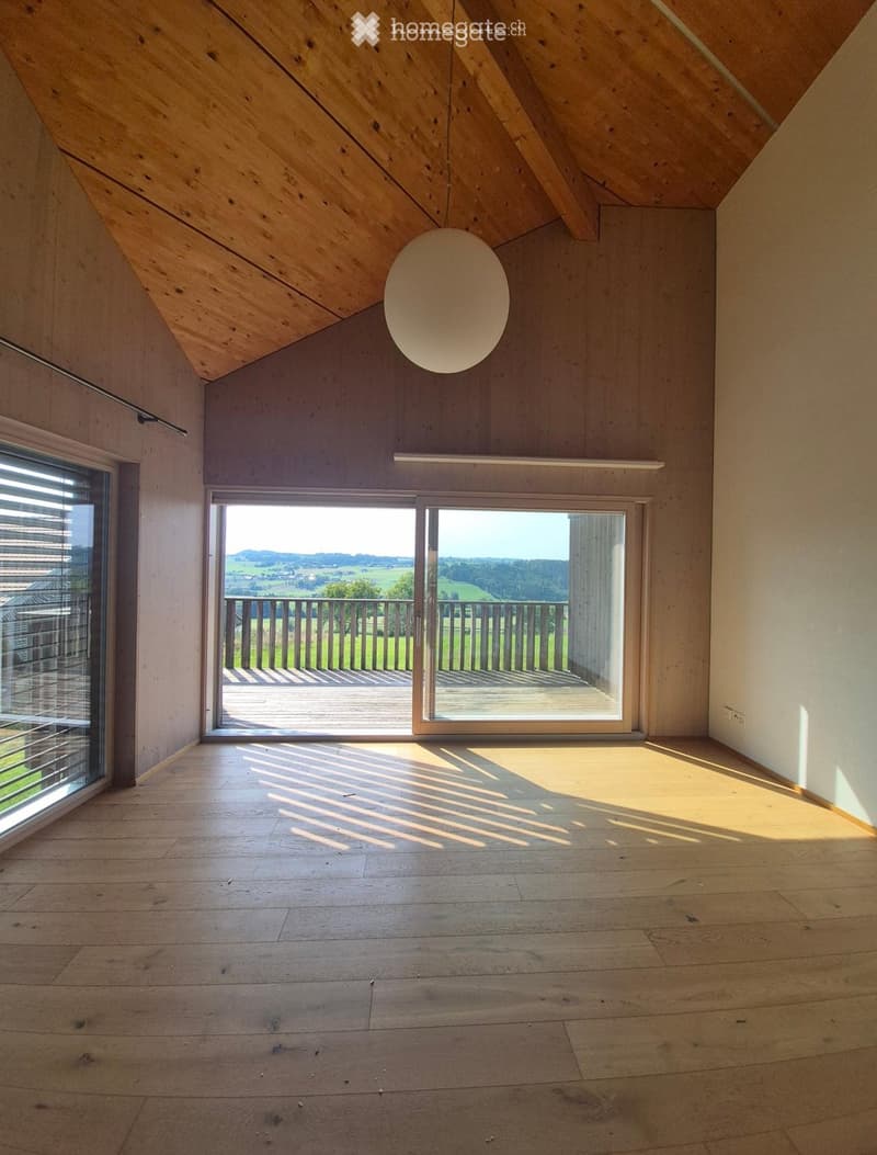 Bright, empty room with wooden flooring and slanted wooden ceiling. Large windows and sliding doors open to a balcony with a scenic view.