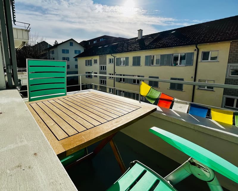 Balcony with wooden table, green chairs, colorful flags, and a view of neighboring buildings.
