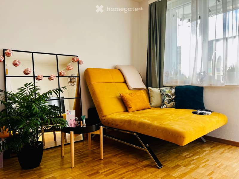 Bright, modern room with a yellow sofa, wooden floor, large window with sheer curtains, and a black side table with a potted plant.