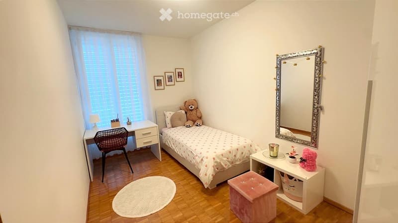 Cozy bedroom with a single bed, desk, chair, mirror, and decorative items including picture frames and teddy bears.