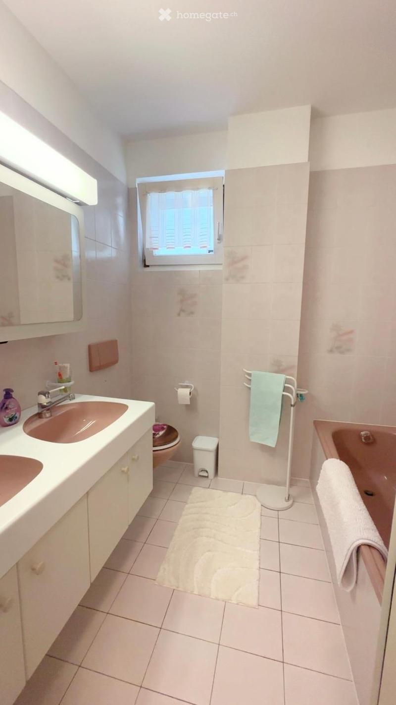 A small, well-lit bathroom with white tiled walls and floor, featuring a double sink vanity, a toilet, a bathtub, a mirror, a towel rack with a towel, and a bath mat on the floor.