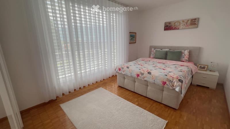 Cozy bedroom with a large bed, floral bedding, nightstand, and a white rug on the wooden floor.