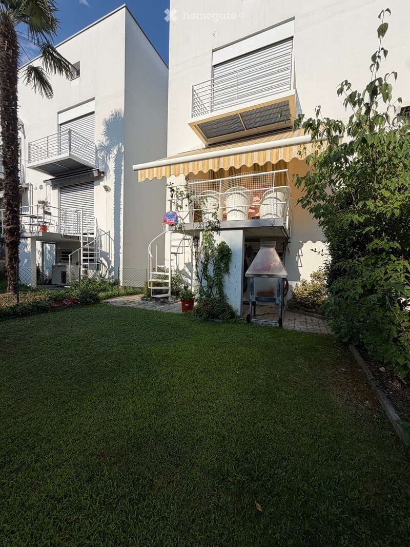 Well-maintained garden with green lawn, patio furniture, barbecue, and a modern white building with balconies and awnings.