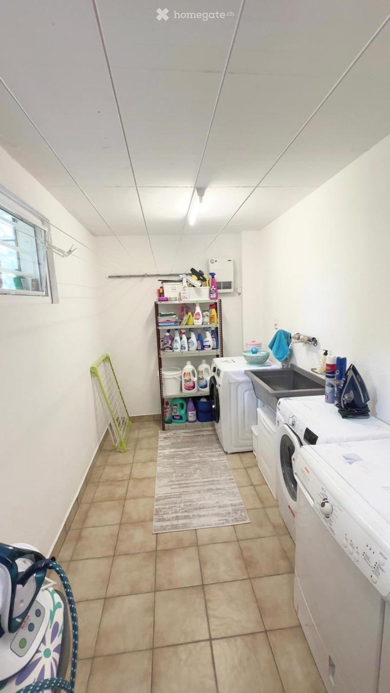 A well-organized laundry room with tiled flooring, white walls, and a window. Features include a washing machine, dryer, sink, ironing board, and shelves stocked with cleaning supplies.