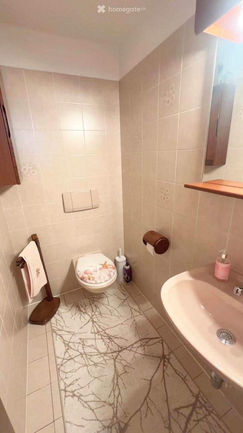 A small, modern bathroom with white tiled walls and a decorative branch-patterned floor. Features include a toilet with a seashell-patterned seat cover, a sink with a pink soap dispenser, a towel on a wooden rack, and a toilet paper holder.