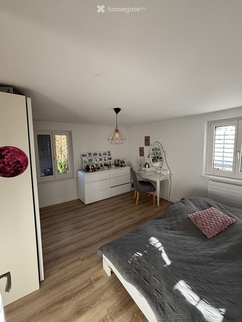 Cozy bedroom with wooden flooring, a grey bed with a pink throw, a white dresser with photos, a vanity table with a mirror, and a hanging pendant light.