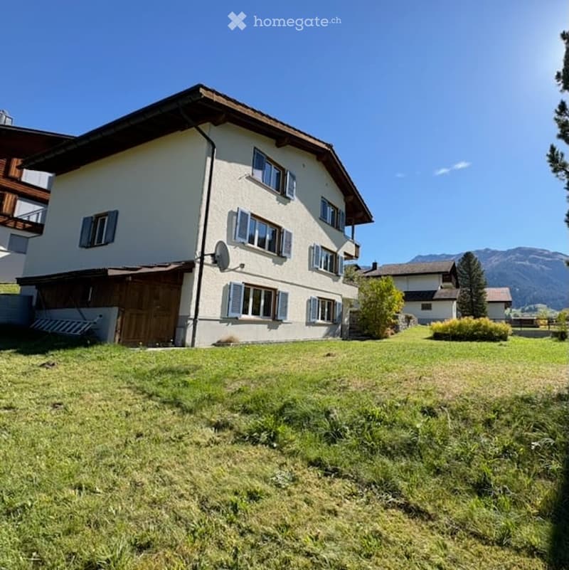 A two-story house with white walls, wooden shutters, and a wooden garage door, situated in a grassy area with a scenic mountain view in the background.