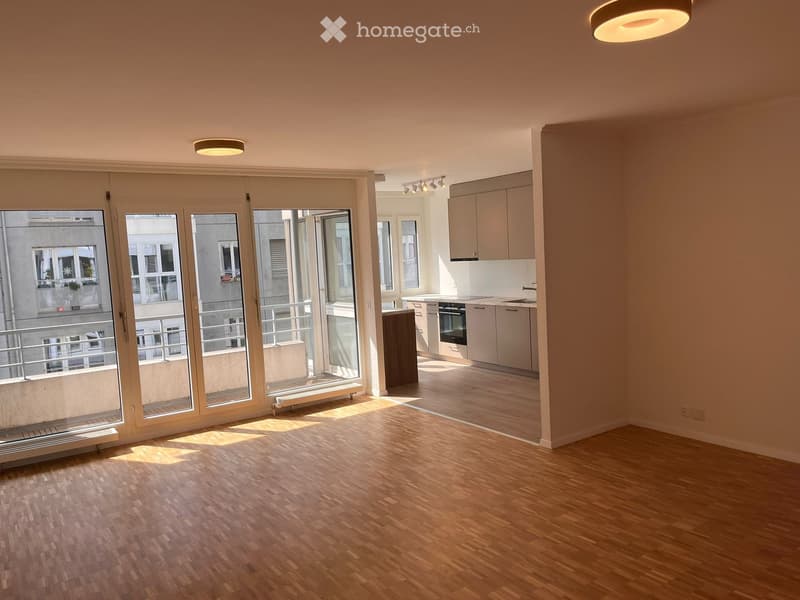 Bright, unfurnished room with wooden flooring, large glass doors leading to a balcony, and a small kitchen area visible in the background.