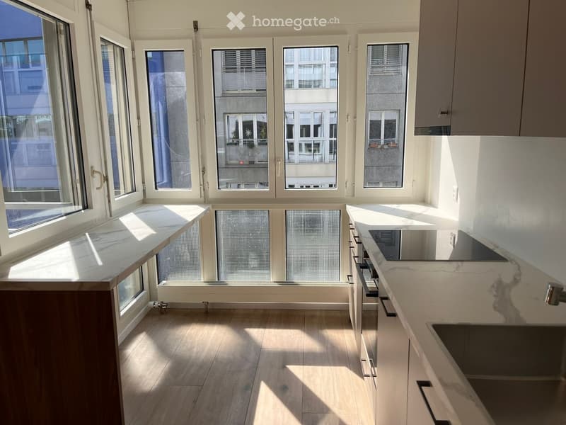 Modern kitchen with light wood flooring, white cabinets, and a marble countertop. Large windows provide ample natural light and a view of the city.