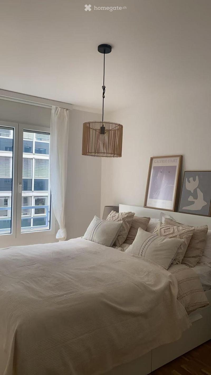 A well-lit bedroom with a large bed, multiple pillows, a textured bedspread, and two framed artworks on the wall. A hanging woven lamp and sheer curtains complete the decor.