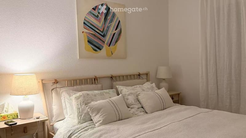 Cozy bedroom with a large bed, multiple pillows, and a colorful leaf-shaped painting on the wall. Two nightstands with lamps flank the bed, and a small table holds a cellphone and a box.