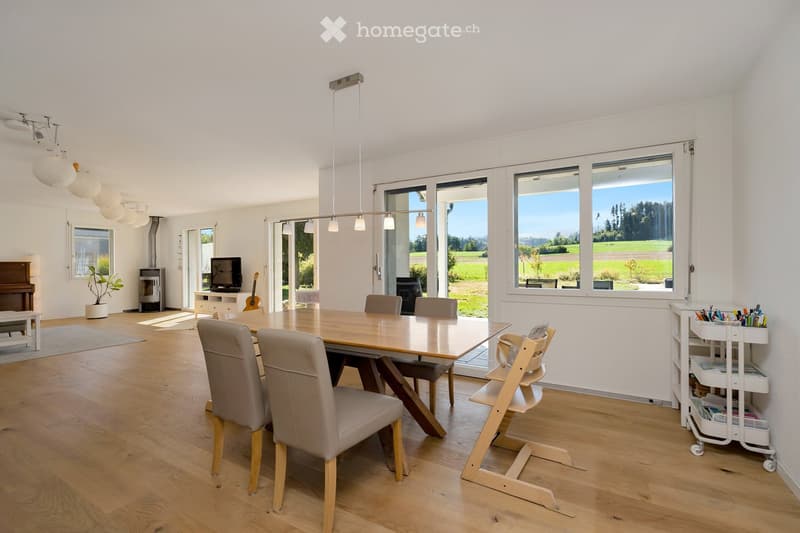 Bright, open dining area with wooden flooring, large windows overlooking a green landscape, a dining table with chairs, a baby chair, a piano, a guitar, a television, a potted plant, and a small cart with supplies.