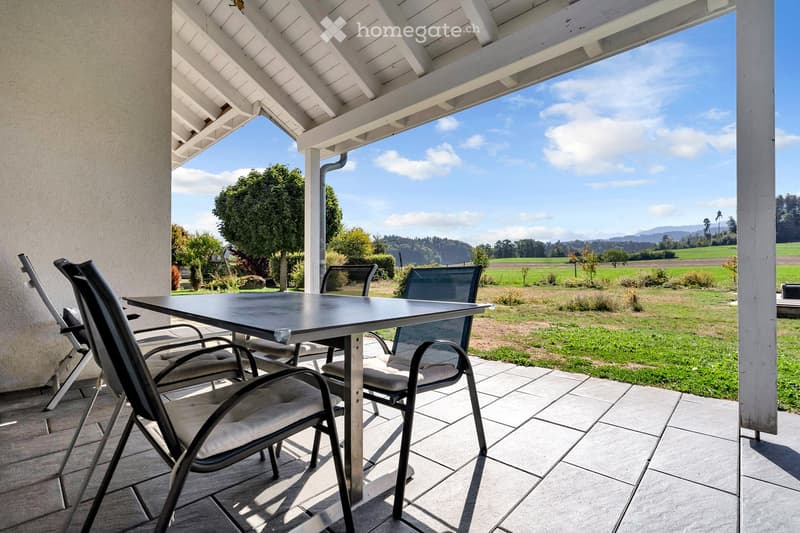A covered patio with a table and chairs, overlooking a grassy field and distant hills under a partly cloudy sky.