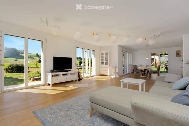 Bright living room with large windows, a grey sofa, coffee table, TV, guitar, and a dining area with a table and chairs.
