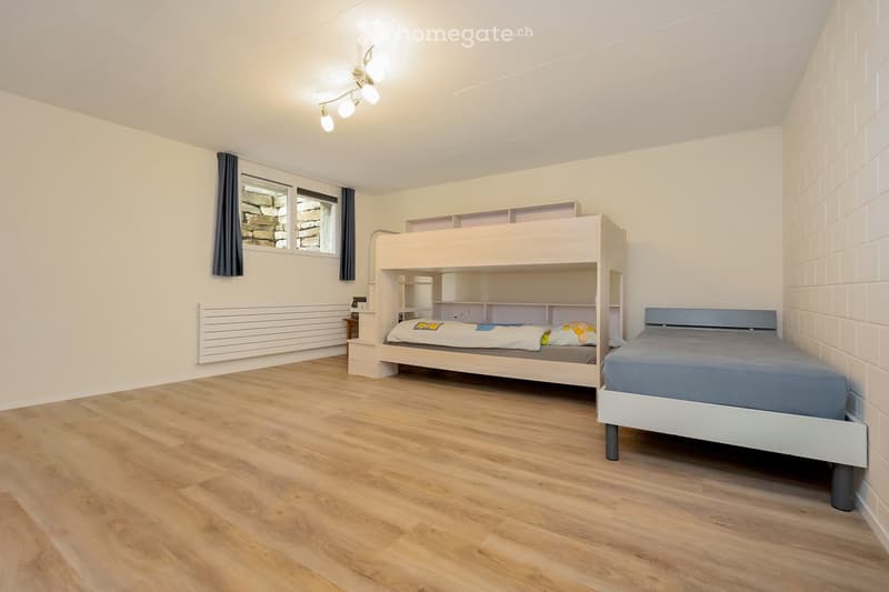 A small bedroom with wooden flooring, featuring a bunk bed and a single bed, both neatly made with bedding. There is a built-in shelf unit next to the bunk bed, a radiator on the wall, and a window with dark curtains.