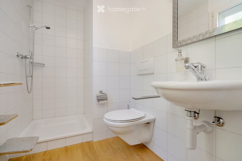 Modern bathroom with white tiled walls, a bathtub, toilet, sink, and a large mirror above the sink. The floor is light wooden, and there are wooden shelves on the left side.