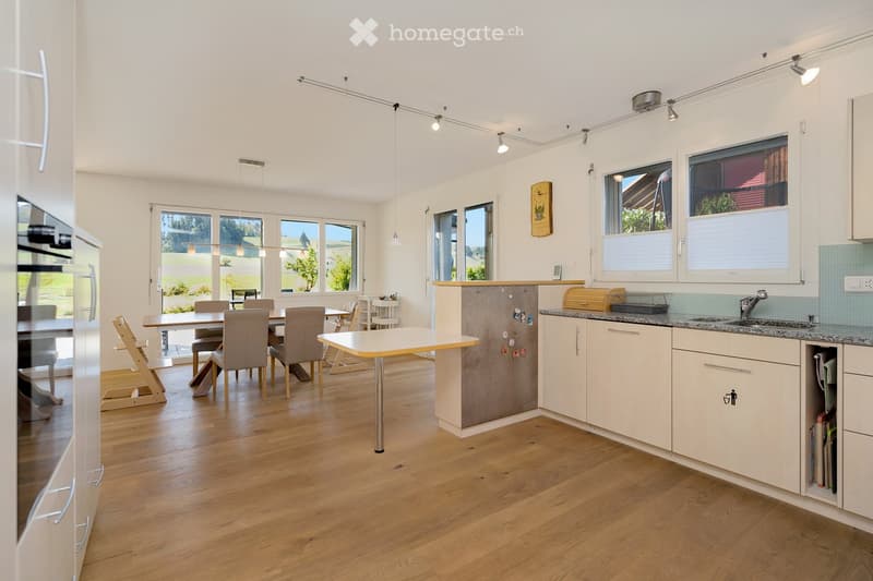 Bright, modern kitchen with white cabinets, granite countertops, and a central island. The space includes a dining area with a table and chairs, a refrigerator, and a sink with a faucet. Large windows provide natural light and a view of the outdoors.