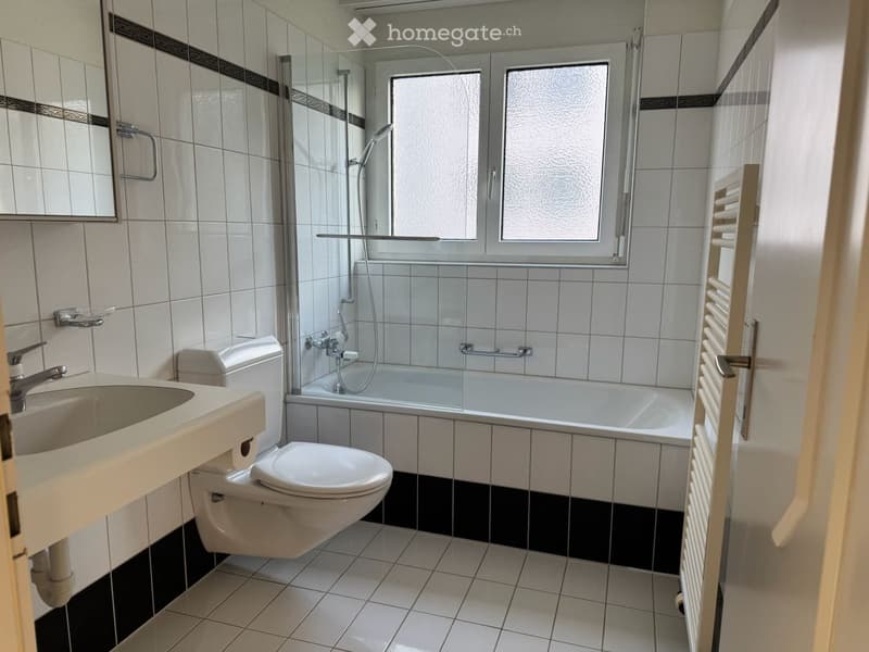Modern bathroom with white tiles, a bathtub with a glass shower enclosure, a toilet, a sink with a faucet, and a radiator.