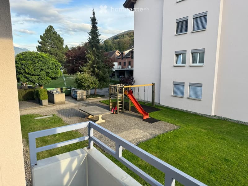 A well-maintained garden area with a small playground featuring a slide, surrounded by a paved pathway and green lawn, with a modern white building in the background and trees in the distance.