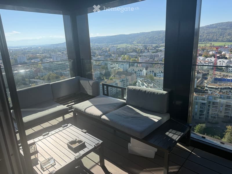 Modern balcony with large windows offering a panoramic city view, featuring a grey sectional sofa and a wooden coffee table.