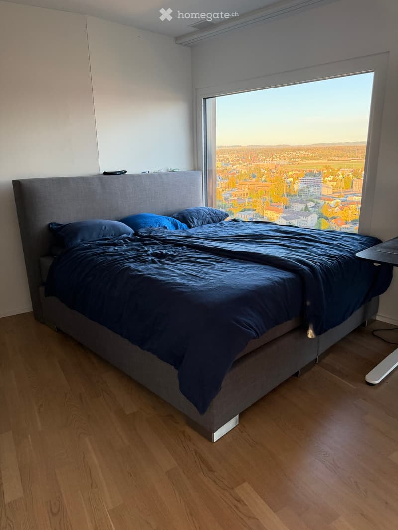 Modern bedroom with a large window offering a scenic city view, featuring a neatly made bed with dark blue bedding and a grey headboard, wooden flooring, and a small table near the window.