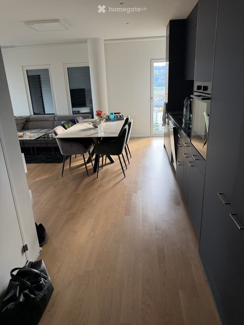 Open-plan living and dining area with light wooden flooring, grey sofa, coffee table, dining table with chairs, and modern kitchen with dark cabinets and stainless steel appliances.