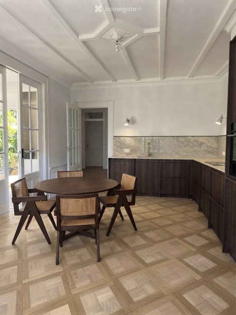 Dining area with a round wooden table and four woven chairs, adjacent to a modern kitchen with dark wooden cabinets and a marble backsplash.