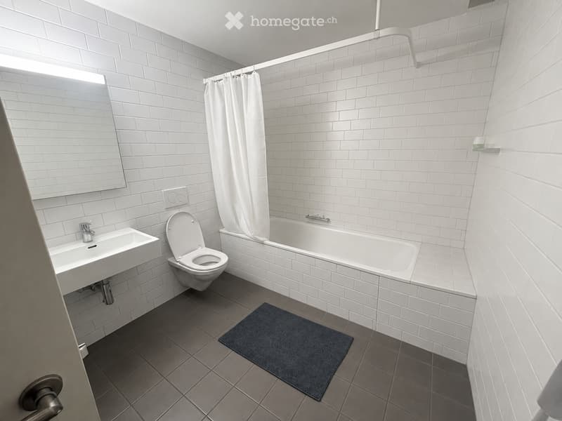 Modern bathroom with white tiled walls and floor, featuring a sink, toilet, bathtub, and a small blue bathmat.