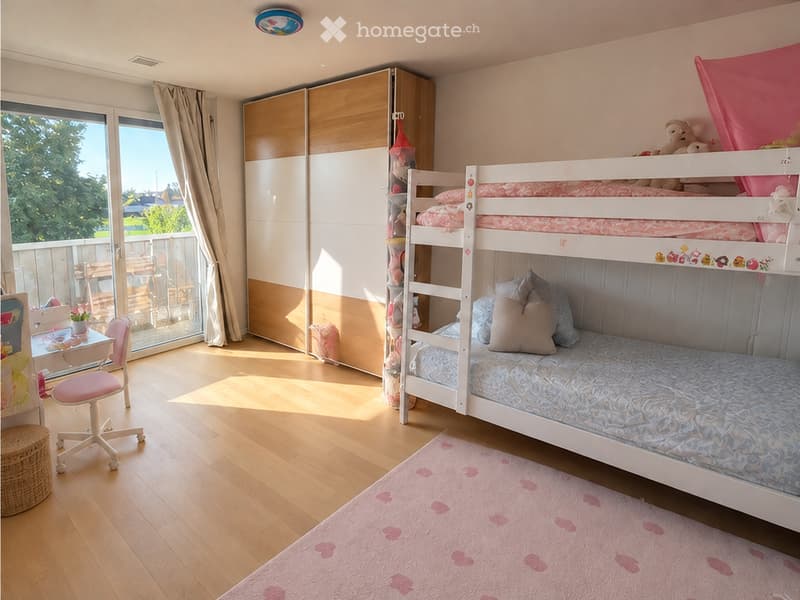 Brightly lit bedroom with a bunk bed, a small desk with a chair, a wardrobe, and a pink rug with heart patterns.