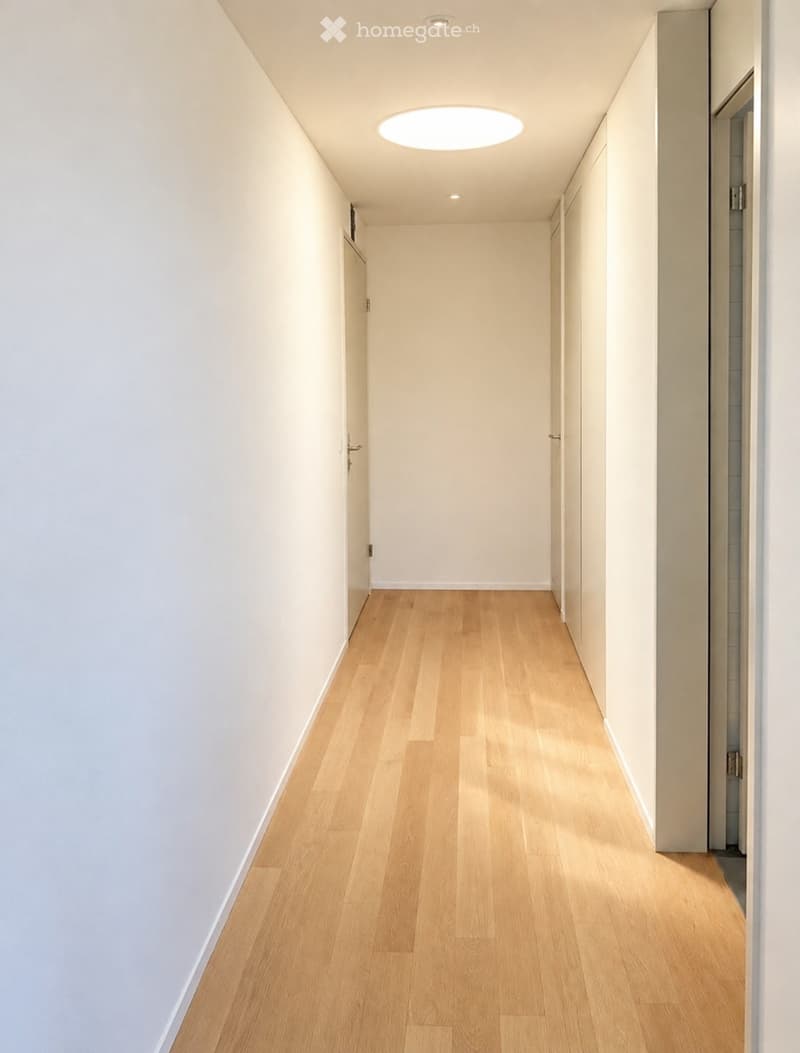 A modern, minimalist hallway with light wooden flooring, white walls, and recessed ceiling lighting. The space is clean and uncluttered, with doors visible on both sides.
