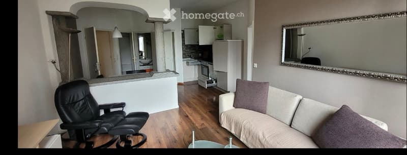 A modern living room with a sofa, a black leather chair, a large mirror, and an open kitchen area with cabinets, a refrigerator, and an oven.