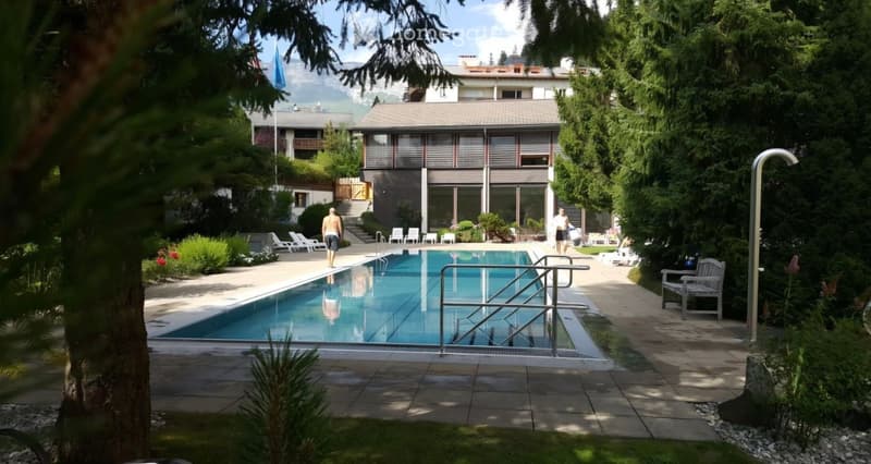 Outdoor area with a swimming pool, surrounded by lounge chairs, greenery, and a building in the background.