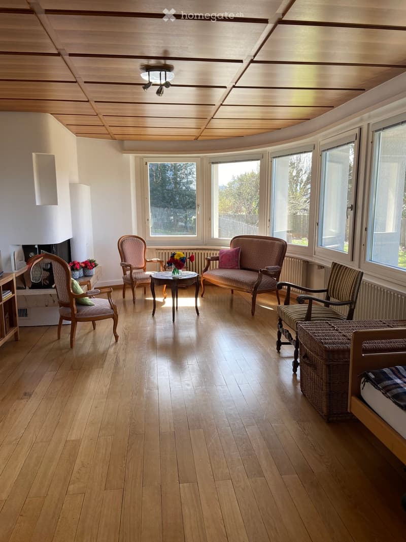 Bright living room with wooden flooring, large windows, and a fireplace. Features a sofa, armchairs, a coffee table with a flower vase, and a wicker chair.