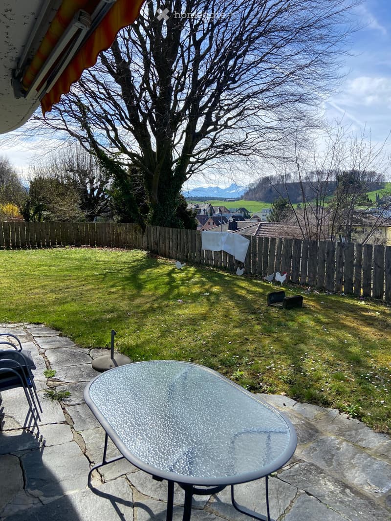 Garden with a round glass table and a chair on a stone patio, surrounded by grass, a wooden fence, and a view of a town and mountains.
