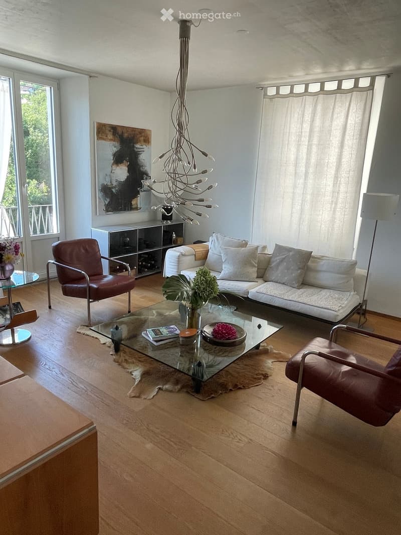 Modern living room with a white sofa, glass coffee table, two leather chairs, a floor lamp, a cabinet, and a chandelier.