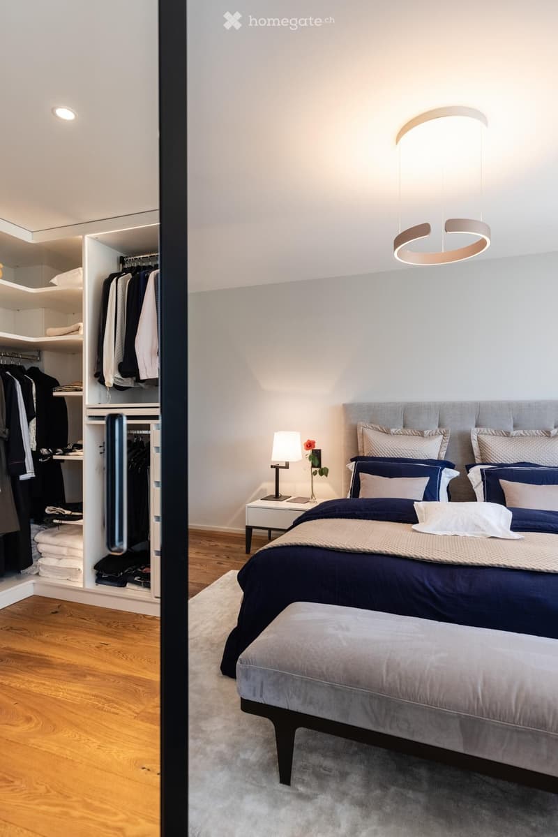 Modern bedroom with a large bed, nightstand, bench, and built-in wardrobe. Features a sleek pendant light and a small flower arrangement on the nightstand.