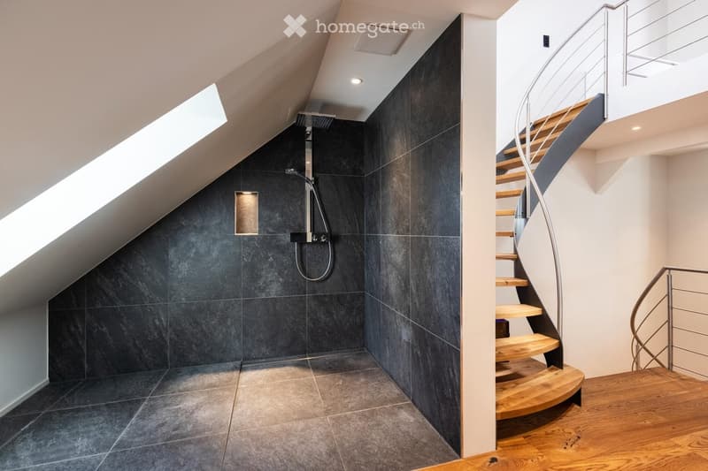 Modern bathroom with dark grey tiles, a sleek shower area, and a wooden spiral staircase leading to an upper level.