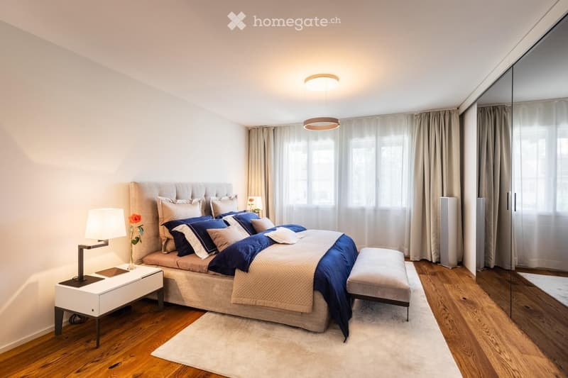 Modern bedroom with a large bed, nightstand, lamp, and a bench at the foot of the bed. The room features wooden flooring, large windows with curtains, and a sleek mirror on the wall.
