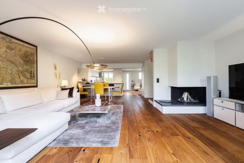 Open-concept living and dining area with white walls, wooden flooring, a white sectional sofa, a coffee table, a fireplace, a television, a dining table with yellow chairs, and a modern kitchen area in the background.