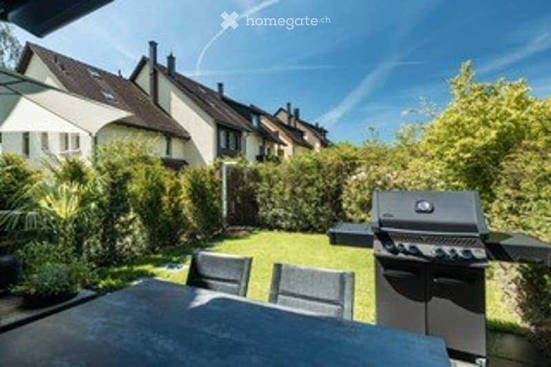 Backyard with a patio table, chairs, and a grill, surrounded by greenery and neighboring houses.