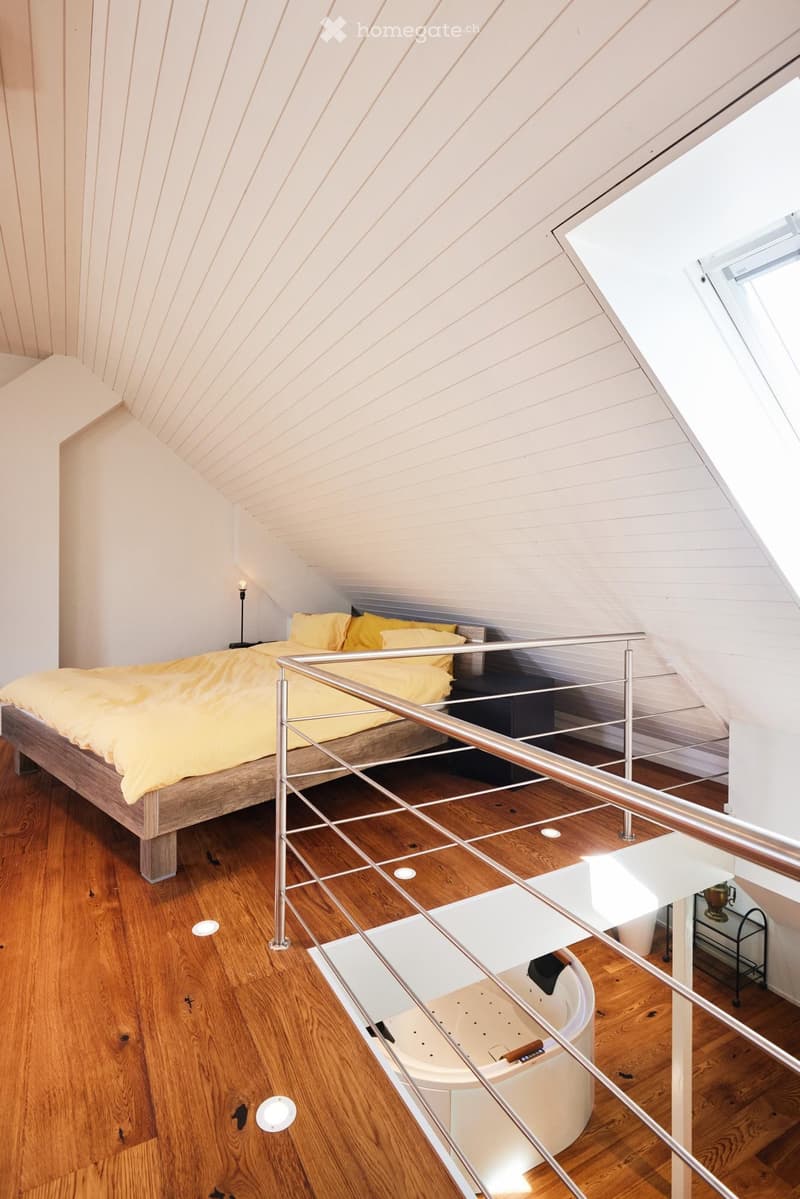 Loft-style bedroom with wooden floors, slanted white ceiling, bed with yellow bedding, nightstand, bathtub, and small table with chair.